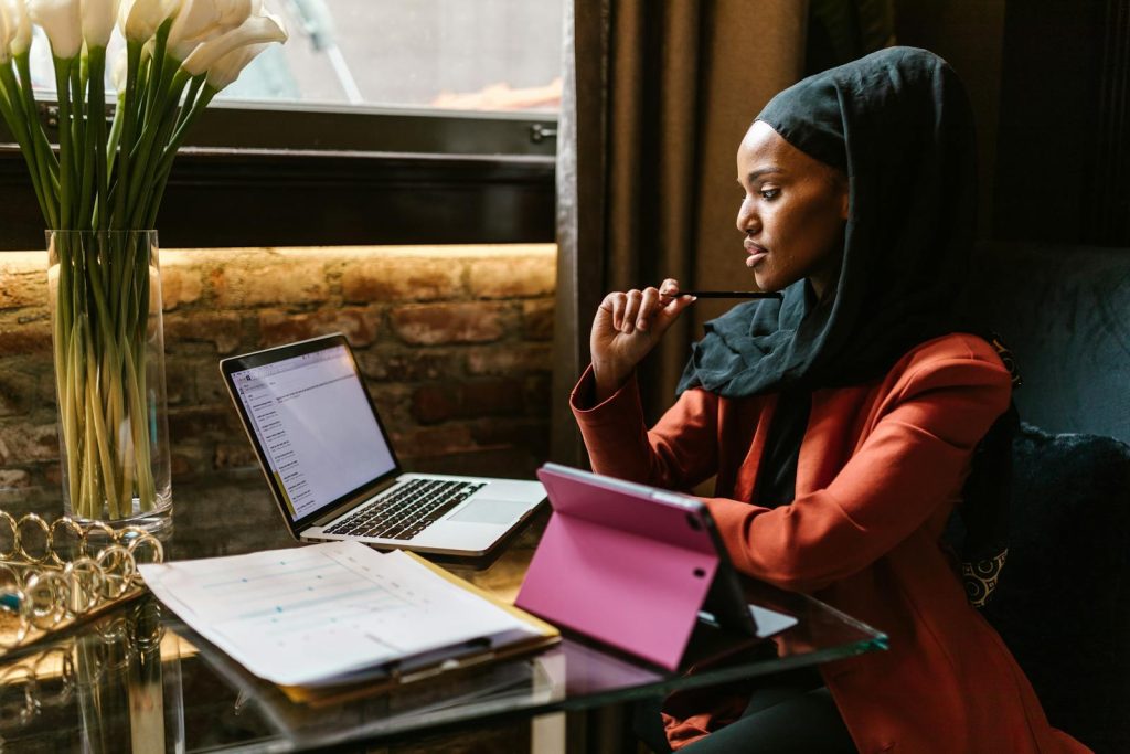 A young Muslim woman in a hijab working with a laptop and tablet indoors, showcasing a modern workspace.